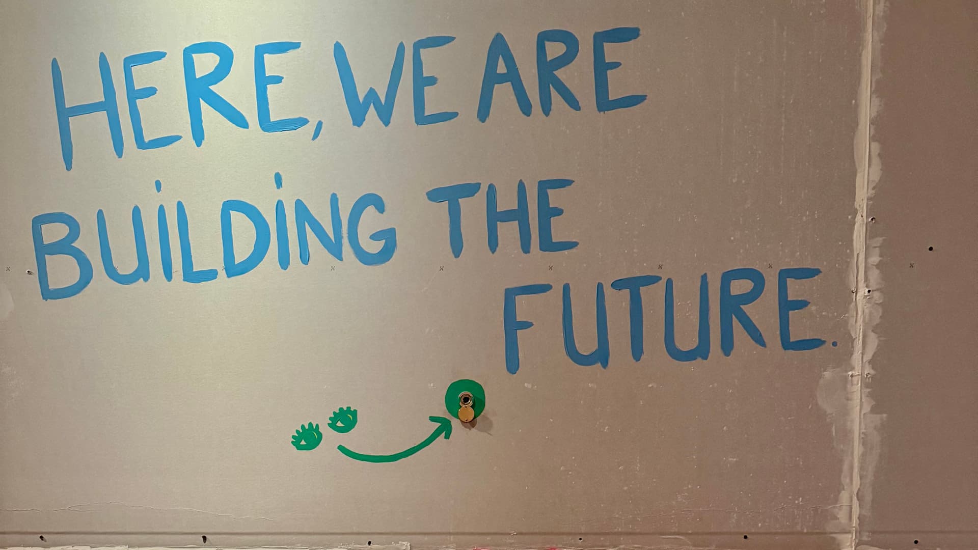 The words “Here we are building the future” are painted in blue on a grey wall; below them, two eyes painted in green and a green arrow indicate the position of a peephole.