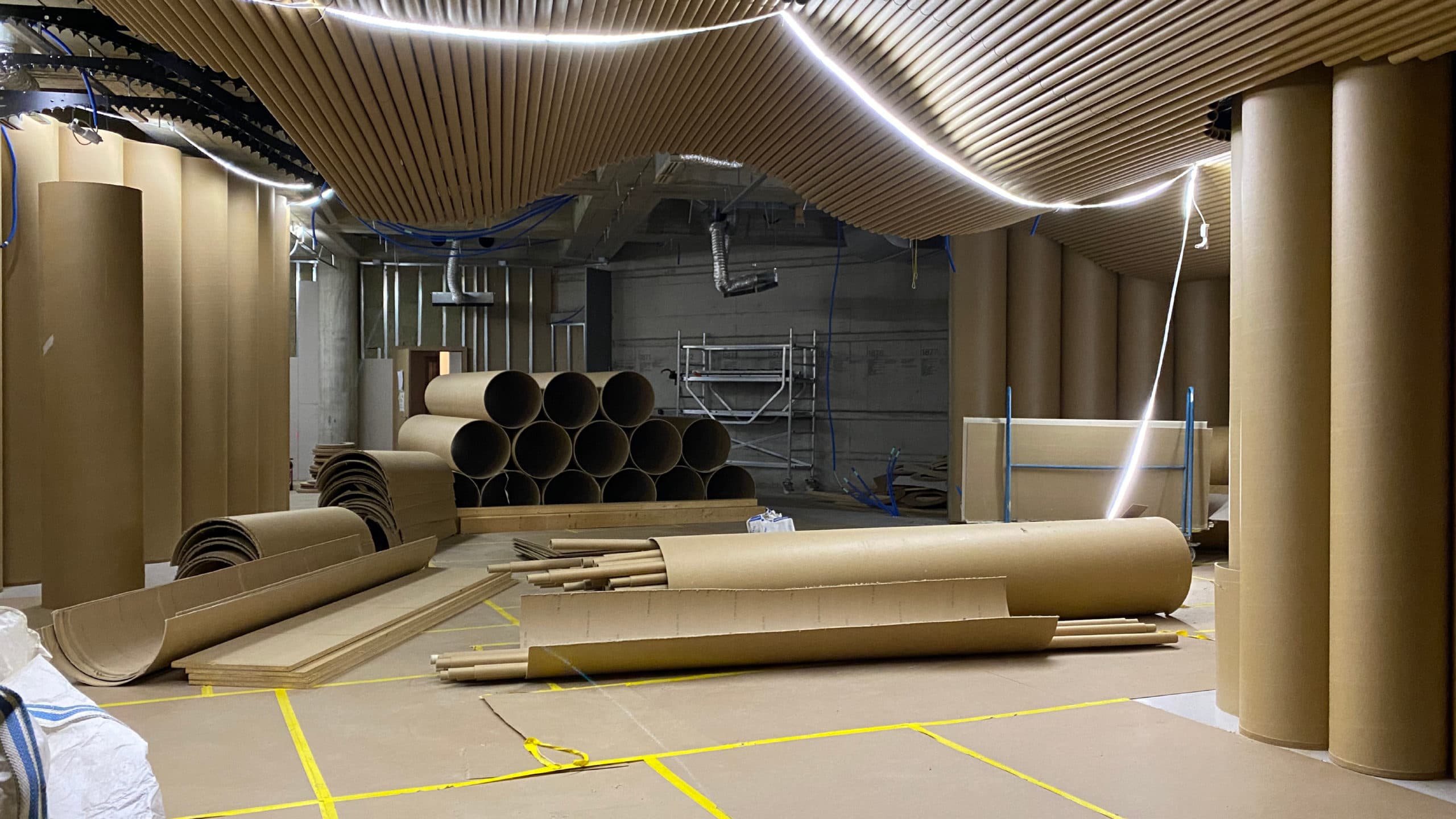 A room undergoing renovation, lit by a powerful string of LED lights, with large cardboard cylinders lining the walls. Some have been dismantled and are piled on top of one another on the floor.