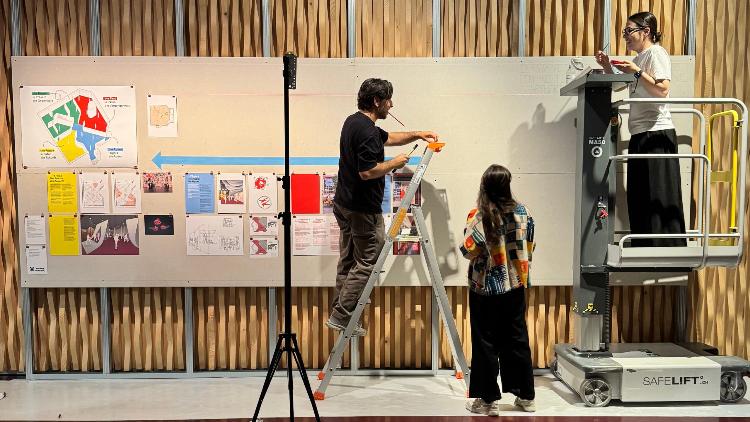 Three people are putting up posters on a large indoor exhibition panel: one is on a ladder, another on a lifting platform and a third is standing on the floor. The panel displays colourful maps, diagrams and documents, set against a wall of wooden slats.