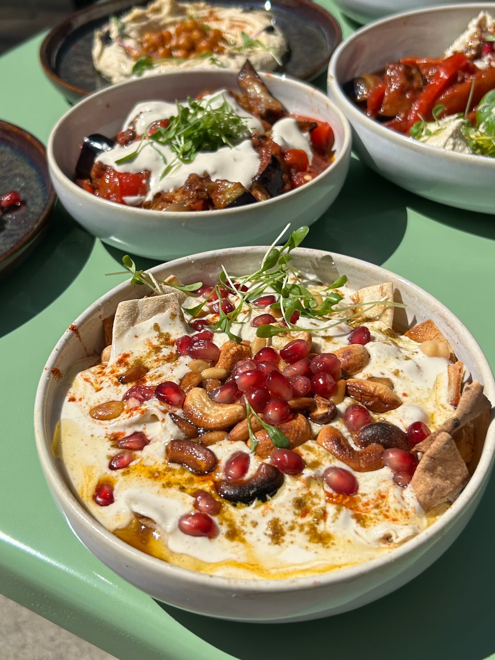 A bowl of hummus topped with chickpeas, cherry tomatoes, fresh herbs and olive oil, placed on a table in the sunshine; other dishes are visible in the background.