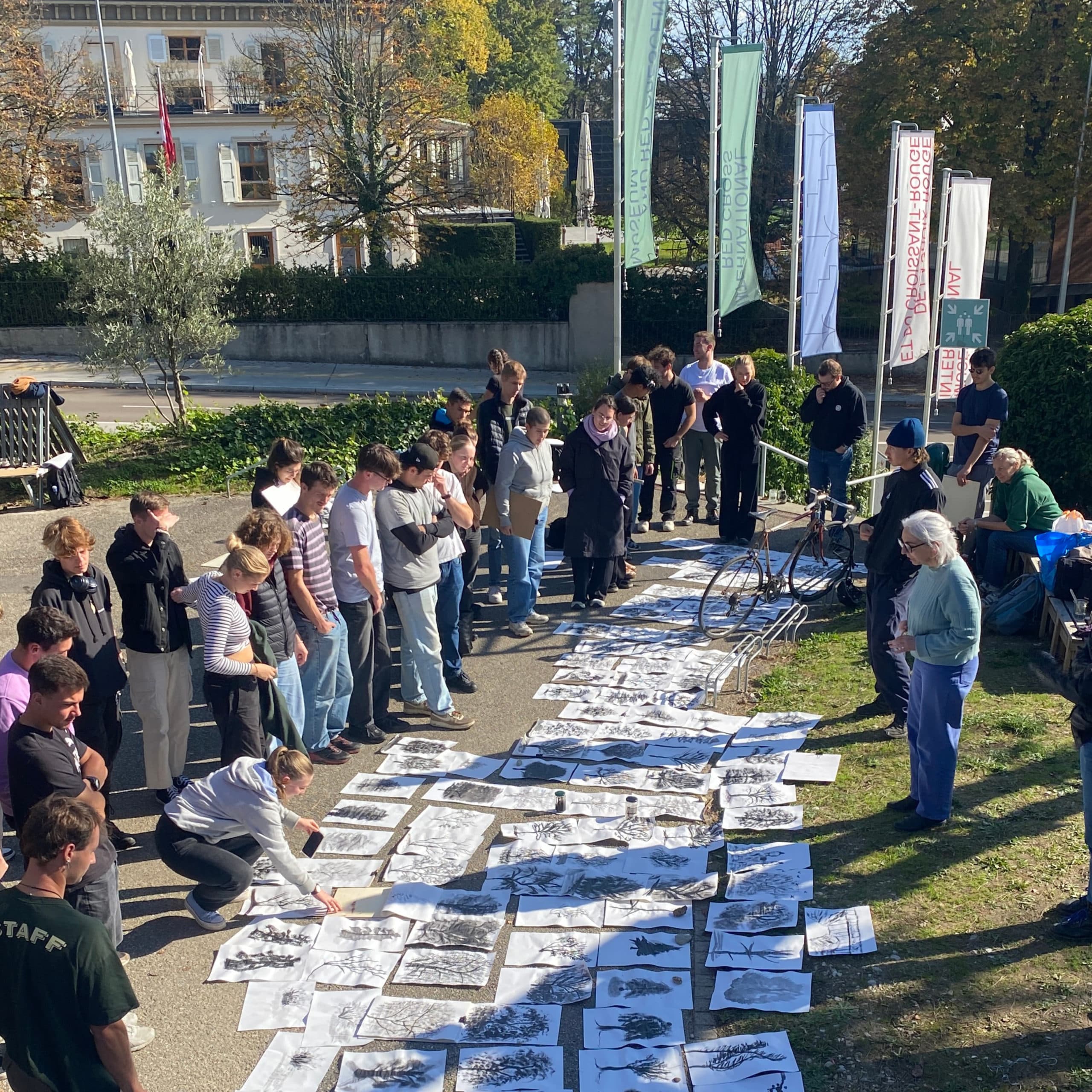 Participants gathered outdoors are examining plans, drawings, and photographs spread out on the ground while one person presents and comments on the documents.