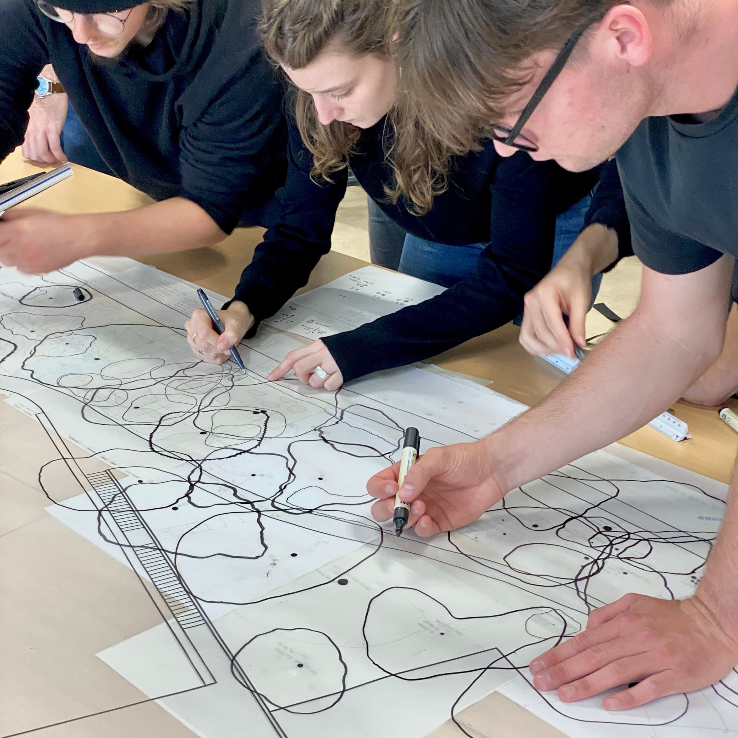 Close-up of several participants working as a team around a table, using markers to draw annotations and shapes on a large printed plan during a collaborative workshop.